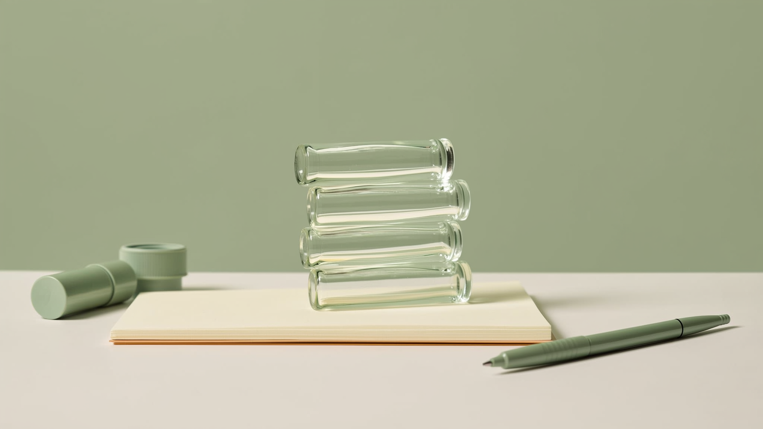 Minimalist still life of glass vials beside a notepad and pen on a clean white desk in cream and sage tones
